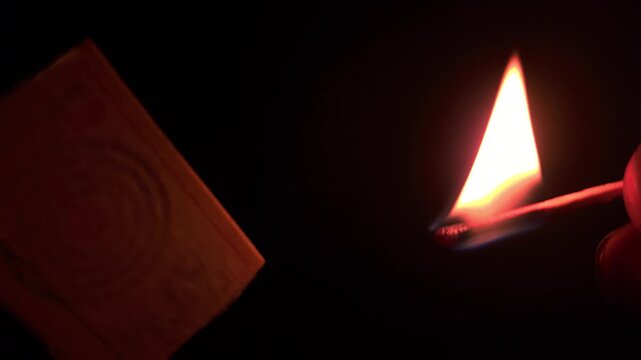 In an extreme close-up macro shot against a black background, fingers strike a sulfur-headed match on the side of the box, and as smoke and fire appear, the flame ignites brightly