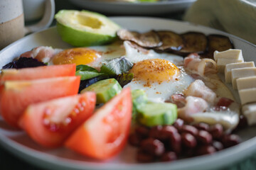 Close-up of a hearty homemade breakfast served on a large plate consisting of fried eggs, bacon, feta cheese, avocado, tomatoes, eggplant, beans, basil leaves.