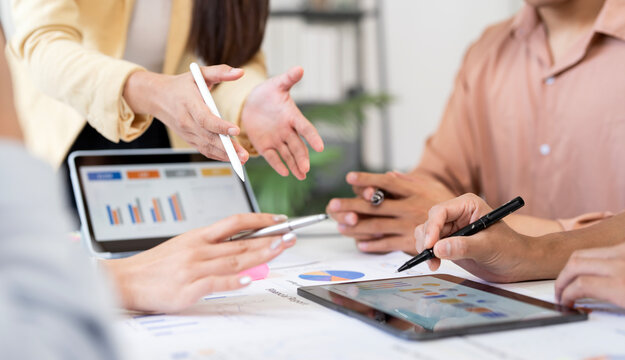 cropped shot of a team of diverse professionals collaborating around a table. They are using a laptop and a tablet with charts and graphs, pointing at the screens and discussing a project.