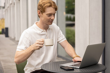 Young Caucasian man sipping tea and reading messages typing on laptop