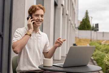 Smiling man multitasking working remotely on laptop and answering client call in open air cafe