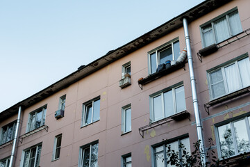 Weathered multi-story residential building with peeling paint, old windows, and visible drainpipes against a clear blue sky, urban housing concept.