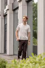 Young ginger male in his twenties walking along the street holding a laptop near modern building with greenery
