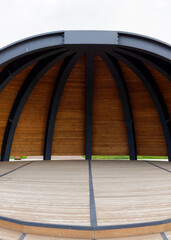 Low-angle view of a contemporary outdoor pavilion stage with curved metal arches, wooden ceiling...