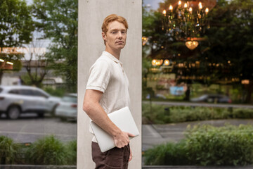 Young male with ginger hair moving along street holding laptop near office building