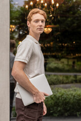 Man walking past office building and passing green flower beds carrying laptop