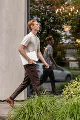 Ginger Caucasian male walking along street holding laptop near office building with greenery