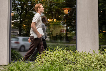 Man walking past office building and passing green flower beds carrying laptop