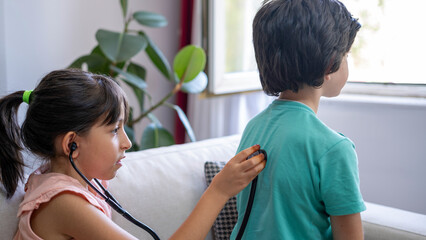 Two siblings are playing doctors. The girl is examining her brother with a stethoscope.