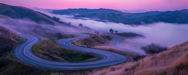 Fototapeta premium A scenic winding road through rolling hills, enveloped in mist, set against a colorful dawn sky.