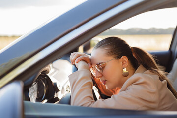 
An exhausted businesswoman with glasses rests her head on her hands on the steering wheel, taking...