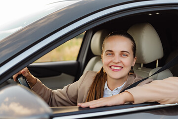 A friendly businesswoman in a beige suit is looking out her car's window and smiling at the camera.