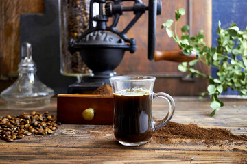 Glass cup of freshly brewed black coffee on a rustic wooden table with ground coffee, beans, and a vintage grinder in the background, styled in cozy farmhouse and artisan coffee photography