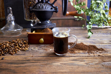 Glass cup of freshly brewed black coffee on a rustic wooden table with ground coffee, beans, and a vintage grinder in the background, styled in cozy farmhouse and artisan coffee photography