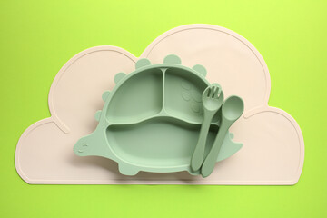 Mint green dinosaur-shaped kids plate with spoon and fork on pastel cloud placemat.