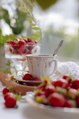 A teacup sits gracefully on a wooden tray surrounded by ripe strawberries and daisies. Bright sunlight illuminates the fresh fruit, creating a vibrant and inviting atmosphere