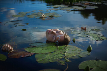 woman lies peacefully in a calm lake, her head resting among green lily pads. Sunlight dances on...