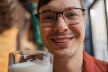 Smiling man in glasses holding a glass of fresh beer while enjoying a refreshing beverage at a...