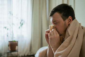 Male patient resting under blanket, experiencing flu symptoms, blowing nose while dealing with...