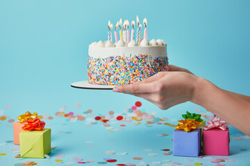 Cropped view of woman holding birthday cake with candles on blue background with confetti and gifts
