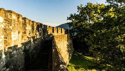 A sun-drenched view of ancient stone castle walls, with lush greenery and a backdrop of distant mountains.