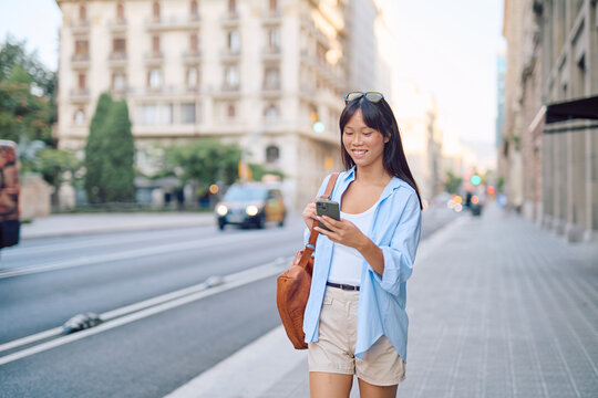 Smiling young woman walking through the vibrant streets of Barcelona, happily using a mobile phone app while enjoying the urban atmosphere
