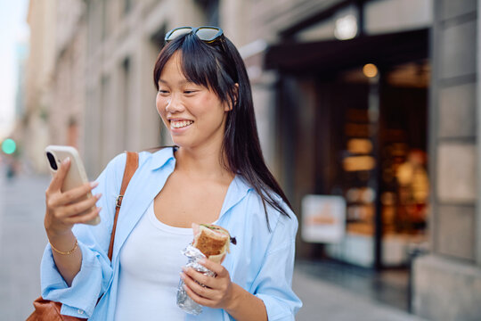 Happy tourist walking through the bustling city center, enjoying a tasty sandwich while using her smartphone to connect with friends