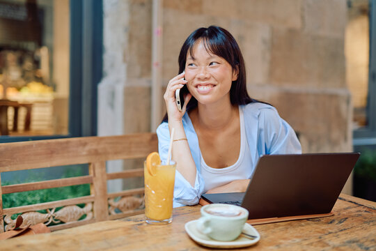 Young asian woman working remotely in cafe, using laptop and talking on smartphone, enjoying orange juice and cappuccino