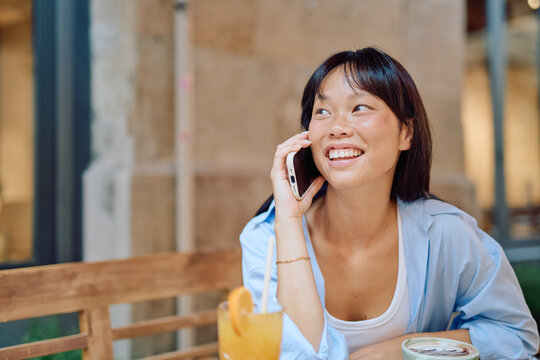 Young woman enjoying a refreshing drink at an outdoor cafe while talking on her phone and smiling - Powered by Adobe