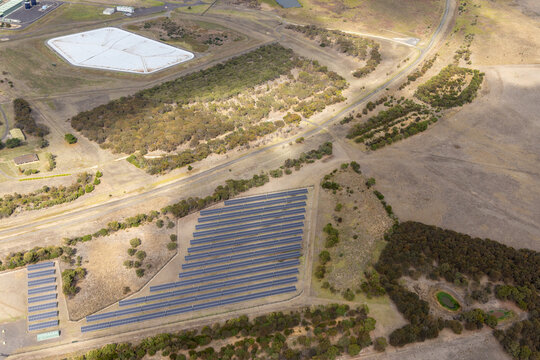 Aerial view of solar panels aligned in rows, reflecting the sky, contrasting with the dry, golden landscape, near a dam, Breamlea, Victoria, Australia.