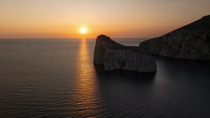 Aerial view of the sun setting over the dark sea and the rocky cliffs of Capo Caccia, creating a mesmerizing contrast of light and shadow, Alghero, Sardinia, Italy.