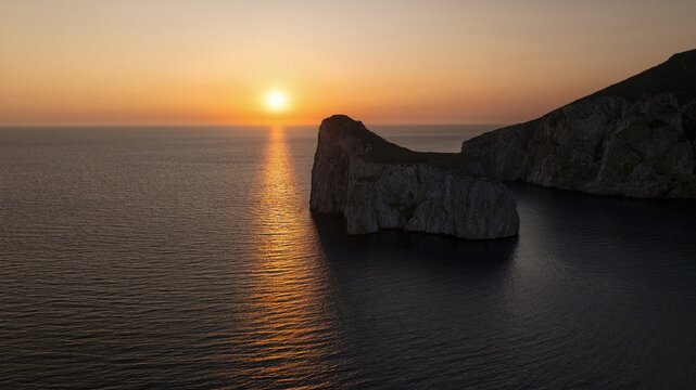 Aerial view of the sun setting over the dark sea and the rocky cliffs of Capo Caccia, creating a mesmerizing contrast of light and shadow, Alghero, Sardinia, Italy. - Powered by Adobe