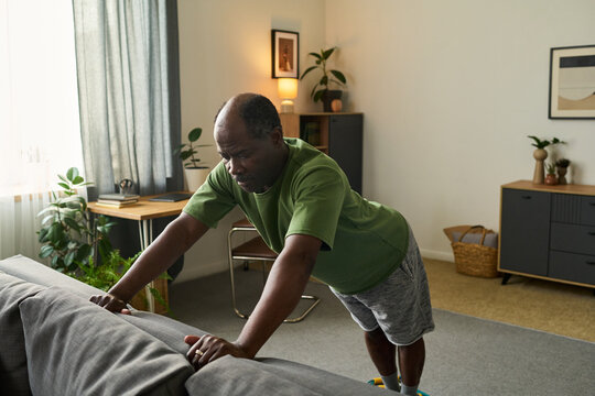 Senior Black man exercising by doing incline push ups against sofa in living room, focusing on fitness and strength training, home environment with plants and decor visible - Powered by Adobe