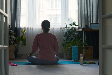 Woman sitting cross legged on yoga mat meditating in living room, back facing camera, surrounded by houseplants and fitness accessories, natural light coming through window