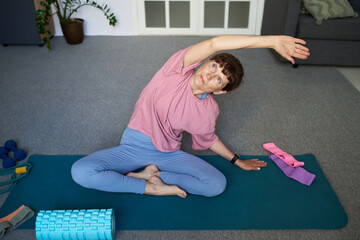 Senior Caucasian woman sitting on yoga mat stretching arm overhead during home workout, exercise...