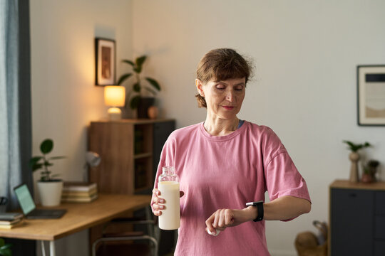 Senior Caucasian woman standing indoors holding bottle of milk while checking smartwatch, appearing focused on time management or healthy lifestyle in home office setting