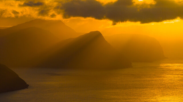 Aerial view of sun rays pierce through the clouds, illuminating the dramatic cliffs and the serene sea of the Northern Isles, Kalsoy, Northern Isles, Faroe Islands.
