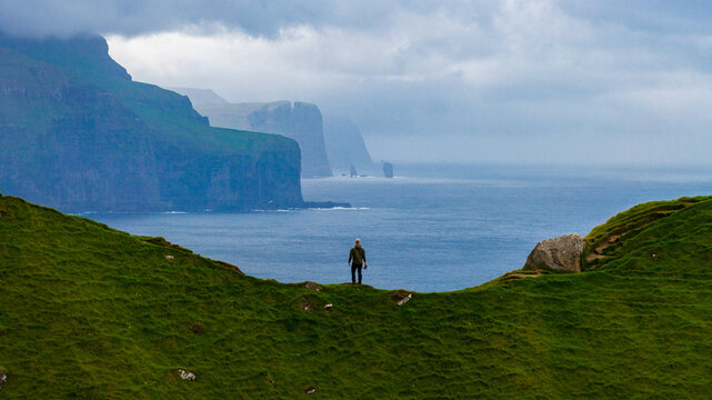 Aerial view of a solitary figure stands atop a vibrant green ridge, gazing out towards the misty, towering cliffs of Kallur, Tr&Atilde;&cedil;llanes, Nor&Atilde;&deg;oya, Faroe Islands.