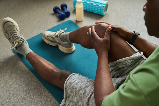 Black Senior man sitting on exercise mat holding knee with both hands, stretching leg muscles after workout, fitness equipment including dumbbells and foam roller visible nearby