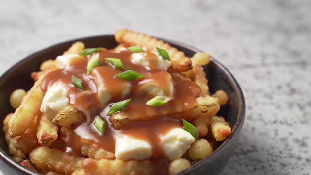 Pouring gravy over poutine with cheese curds and fries in a dark bowl on a light surface view