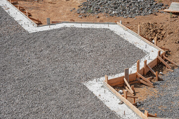 A newly poured concrete slab foundation and pathway, framed by wooden formwork and surrounded by loose gravel and brown soil on a sunny construction site.