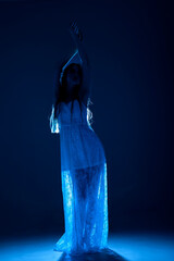 Woman in flowing white dress poses gracefully in a dark blue illuminated setting during a creative performance