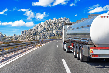 A fuel tanker truck is driving on a highway through a mountainous landscape under a bright blue sky.
