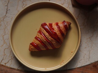 A freshly baked croissant with red stripes served on a beige plate, accompanied by a cup of...