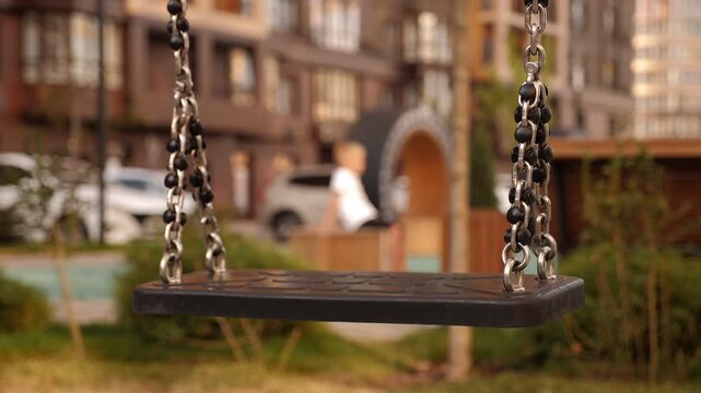 A sad little boy leaves the playground alone, in the foreground of a swing on chains. A modern playground with an empty swing.