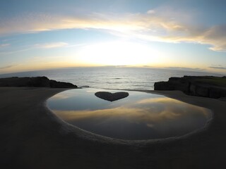 Heart shaped pool reflecting the sky and ocean at sunset with rocky shoreline in the distance