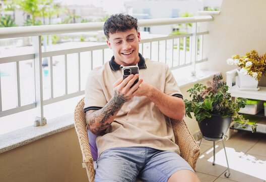Young caucasian smiling 20 years old boy talking on his smartphone sitting in outdoor terrace enjoying tech and social. Modern lifestyle, relaxation, technology use - Powered by Adobe