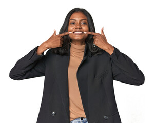 Young Indian woman in business suit smiles, pointing fingers at mouth.
