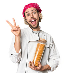 Young chef holds pasta sauce jar in studio joyful and carefree showing a peace symbol with fingers.
