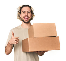 Young man with cardboard boxes in studio smiling and raising thumb up